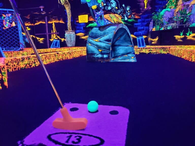 A monster mini golf putter next to a neon golf ball on a glow-in-the-dark indoor golf course.